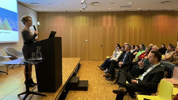 DiSCussion: What can AI do? Marie-Luisa Frick, photographed from the side, stands at the lectern, microphone in hand, and gives a lecture.