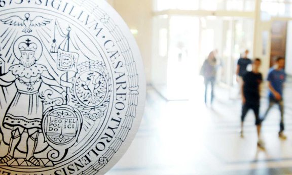 Im Vordergrund das Siegel der Universität Innsbruck, im Hintergrund gehen Menschen in Bewegungsunschärfe durch das Hauptgebäude
