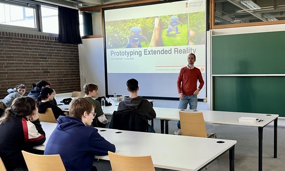 Auf dem Bild zu sehen sind Schülerinnen und Schüler an Tischen in einem Hörsaal. Im Zentrum des Bildes ist eine Leinwand zu sehen mit einer Präsentationsfolie zum Workshop "Prototyping Extended Reality". Vor der Leinwand steht Pascal Knierim.